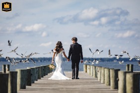 At Herrington on the Bay, North Beach, MD, the bride and groom walk along the wooden pier just after their ceremony, sending gulls scattering into the sky as they enjoy their beautiful waterfront surroundings.